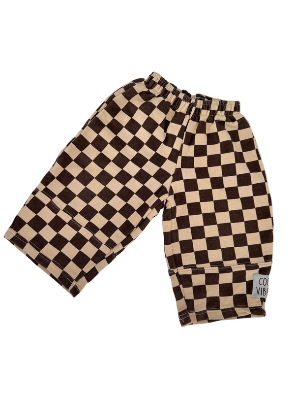 A pair of shorts with a brown and beige checkerboard pattern, laid flat on a white background with soft leaf shadows.