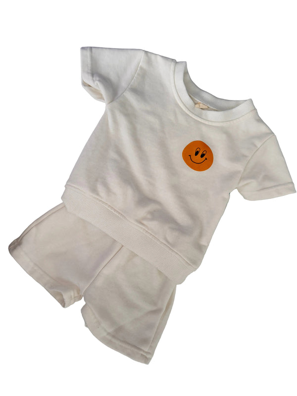 A white two-piece baby outfit set consisting of a short-sleeve top and matching shorts, featuring a small orange smiley face graphic on the chest, laid flat on a light gray textured surface.