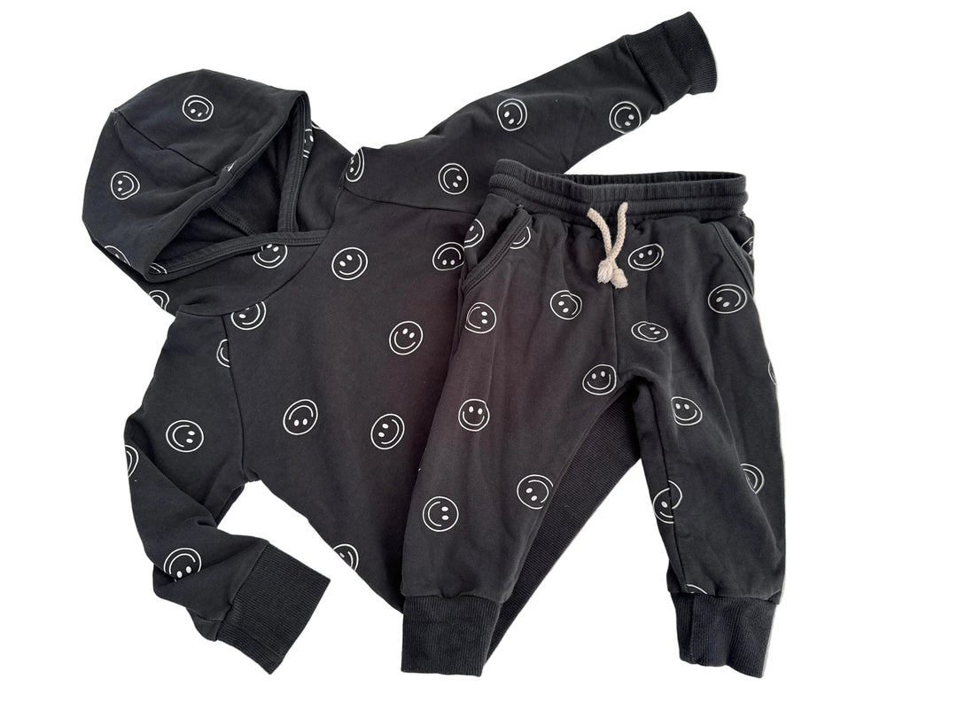 A children's dark gray hooded sweatshirt and matching jogger pants set, both featuring an all-over white smiley face pattern. The hoodie has a hood and long sleeves, while the joggers have an elastic waistband with a white drawstring.