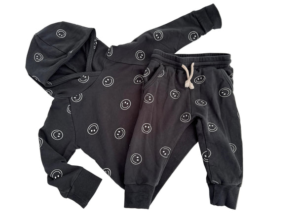 A children's dark gray hooded sweatshirt and matching jogger pants set, both featuring an all-over white smiley face pattern. The hoodie has a hood and long sleeves, while the joggers have an elastic waistband with a white drawstring.