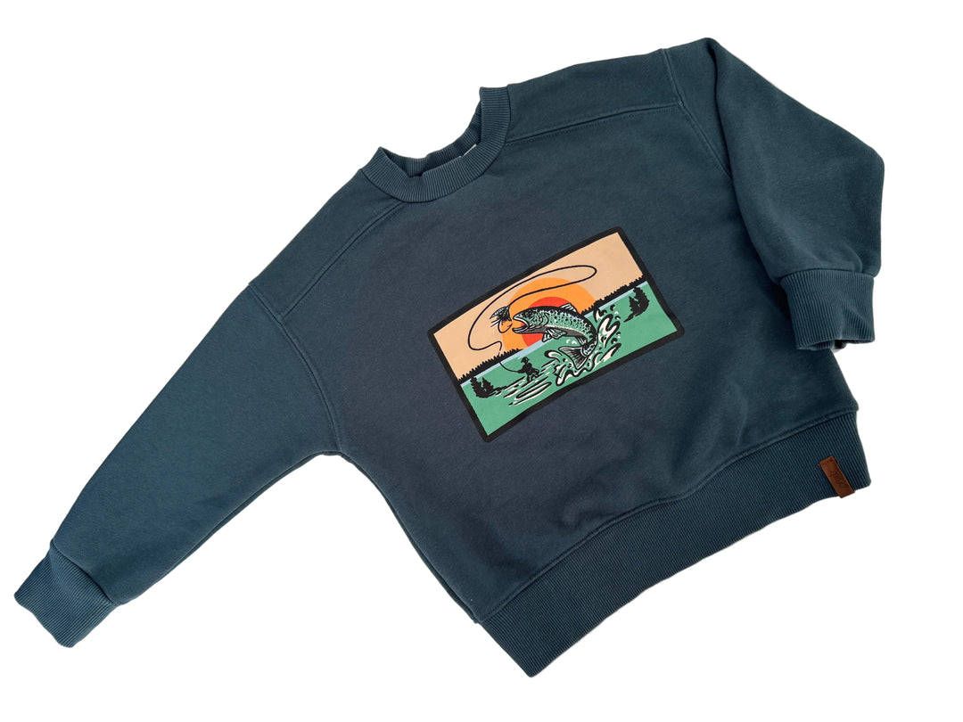 A dark slate blue crewneck sweatshirt with a rectangular graphic patch on the chest showing a stylized fish in green, orange, and beige waves. The sweatshirt is laid flat on a white surface with soft shadows.
