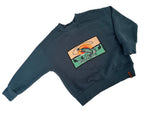 Load image into Gallery viewer, A dark slate blue crewneck sweatshirt with a rectangular graphic patch on the chest showing a stylized fish in green, orange, and beige waves. The sweatshirt is laid flat on a white surface with soft shadows.
