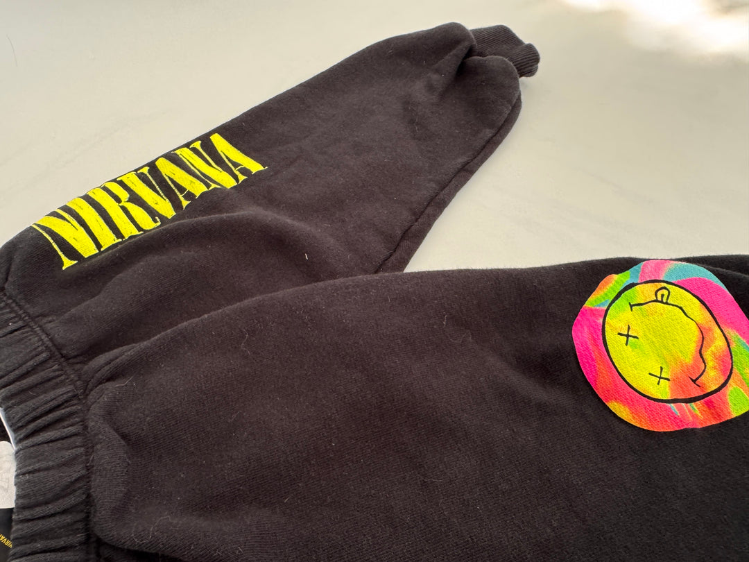 Black sweatpants featuring a bright yellow 'NIRVANA' logo printed on one leg and a colorful neon smiley face patch on the other, laid flat on a light surface.