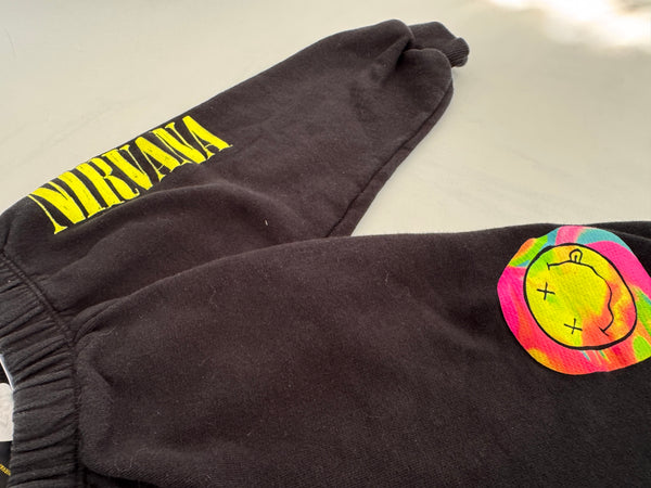 Black sweatpants featuring a bright yellow 'NIRVANA' logo printed on one leg and a colorful neon smiley face patch on the other, laid flat on a light surface.