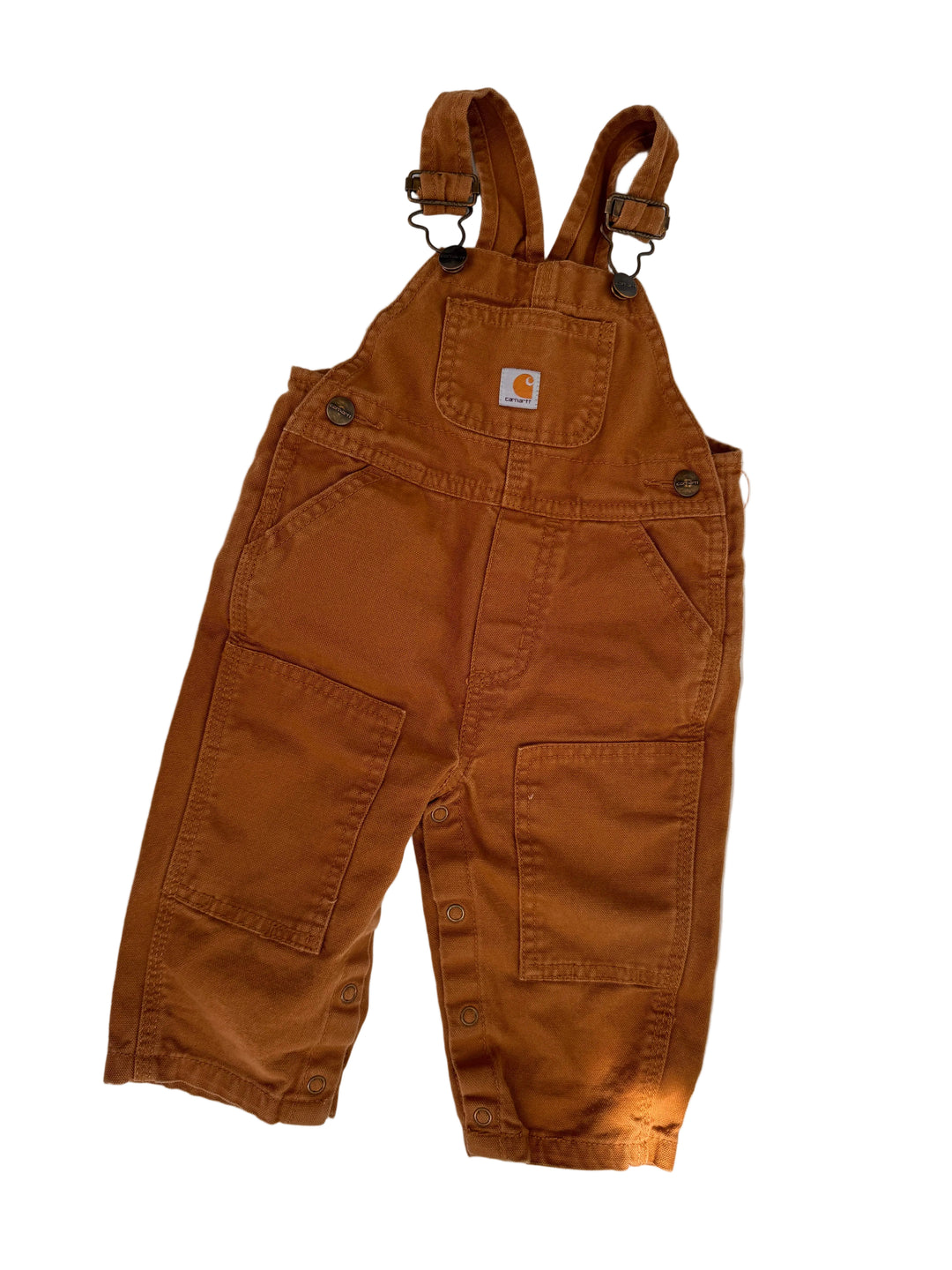 A pair of brown Carhartt bib overalls laid flat on a light gray surface with soft leaf shadows. The overalls feature a classic Carhartt logo patch on the chest pocket, metal snap closures, and adjustable shoulder straps.