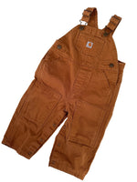 Load image into Gallery viewer, A pair of brown Carhartt bib overalls laid flat on a white background, featuring adjustable shoulder straps, front patch pockets, and button details.
