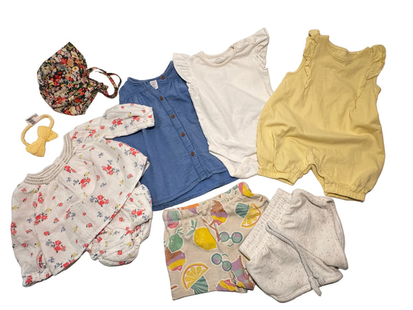 A collection of baby clothing items laid out on a white surface, including a yellow sleeveless romper, a white short-sleeve top, a blue button-front top, a floral long-sleeve romper, a floral hat, a yellow pacifier clip, and patterned shorts.
