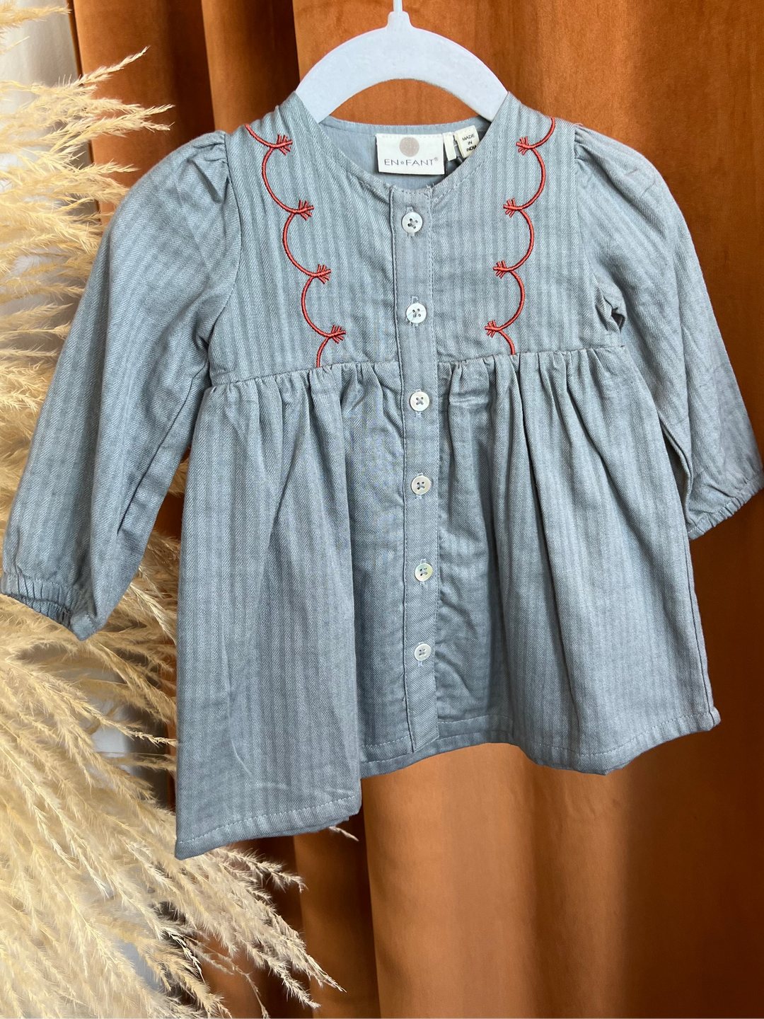 A light blue-gray striped long-sleeve dress for a toddler girl, featuring red embroidered scalloped lines along the neckline and a button-up front, hanging on a white hanger against a warm brown curtain backdrop.