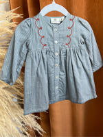 Load image into Gallery viewer, A light blue-gray striped long-sleeve dress for a toddler girl, featuring red embroidered scalloped lines along the neckline and a button-up front, hanging on a white hanger against a warm brown curtain backdrop.
