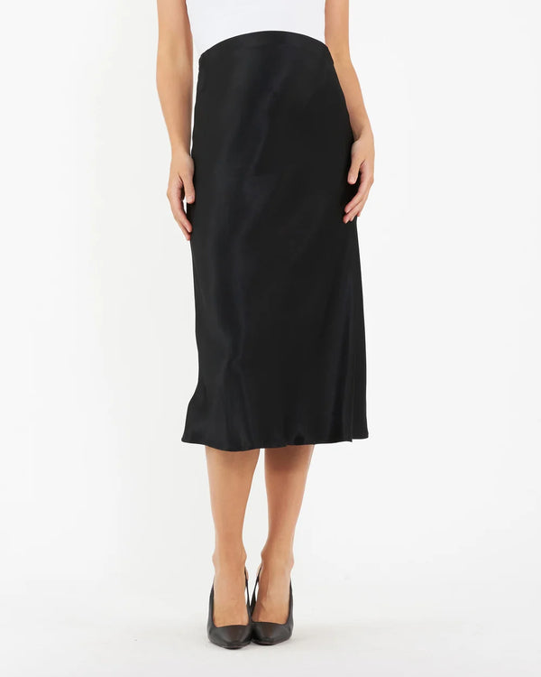 A model wearing a black satin midi skirt with a subtle sheen, paired with a white top and black pointed-toe heels, shown against a plain white studio background.